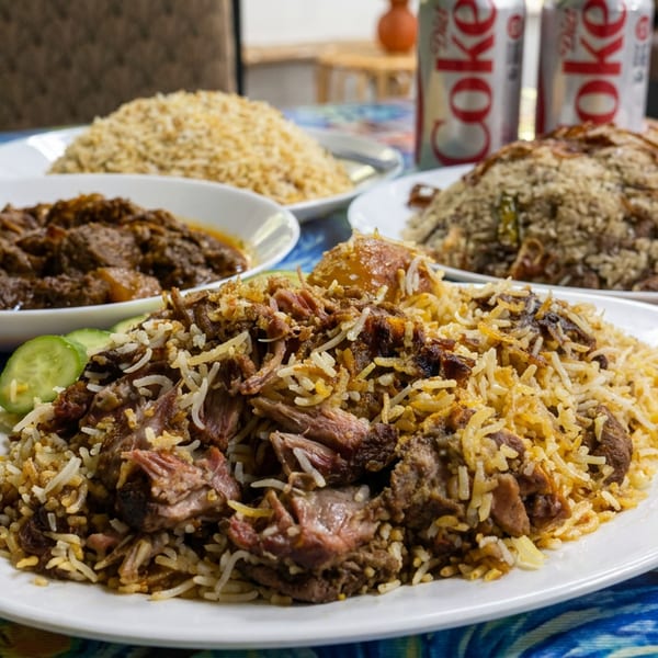 Platters of spiced rice with tender meat, stewed meat, and rice dishes arranged on a colorful restaurant table beside Coke cans.